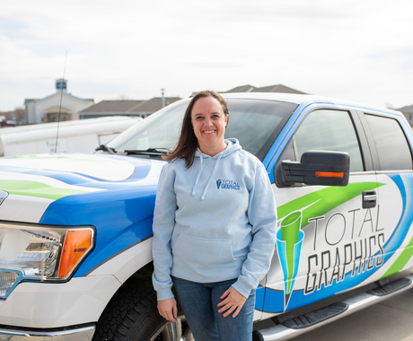 White woman with shoulder length brown hair wearing a light blue hoodie sweatshirt stands in front of a truck with blue, white, and green designs on it and words 'Total Graphics' on the driver side 
